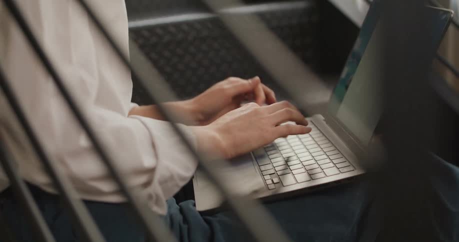 A woman with light hair sits on dark metal stairs inside a building looks downward in thought opens a silver computer begins typing on the keyboard and works framed by diagonal black railings