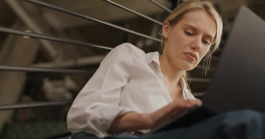 A focused blond woman wearing a white shirt and jeans sits on a staircase working intently on a black laptop with her head occasionally moving as she thinks