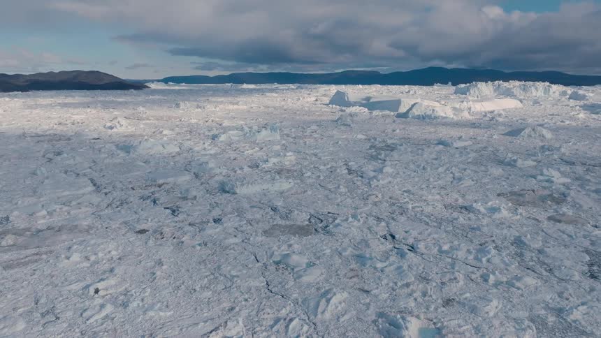 An aerial view of the frozen arctic sea shows a vast field of broken ice floes extending to large icebergs and distant dark mountains under a cloudy sky