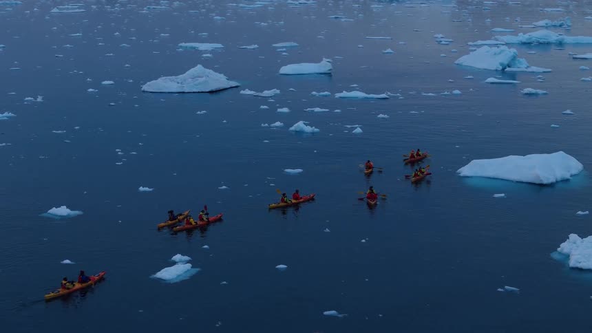 An aerial view shows a group of kayakers paddling together through a vast calm sea dotted with numerous icebergs and ice floes