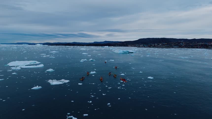 Aerial wide shot of orange kayaks among ice floes near a coastal town