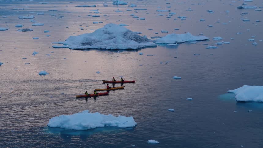 Aerial view of people paddling kayaks through icy waters with icebergs