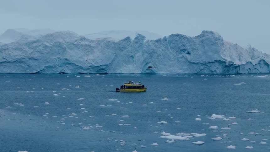 A small yellow boat navigates the dark blue arctic ocean surrounded by floating ice chunks with a giant fractured blue glacier cliff rising dramatically in the foggy background
