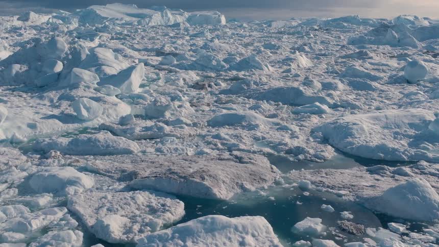 An expansive aerial view shows a lone person in a red jacket standing on a rocky outcrop observing a vast fjord filled with countless chunks of floating white and blue ice