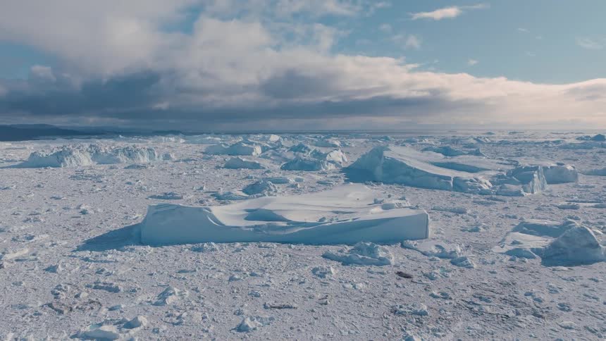 A vast plain of fractured sea ice stretches toward dark hills beneath heavy clouds