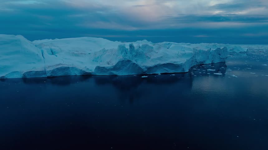 The camera flies over dark icy waters toward and across a massive craggy blue tinted iceberg under a dramatic cloudy sky