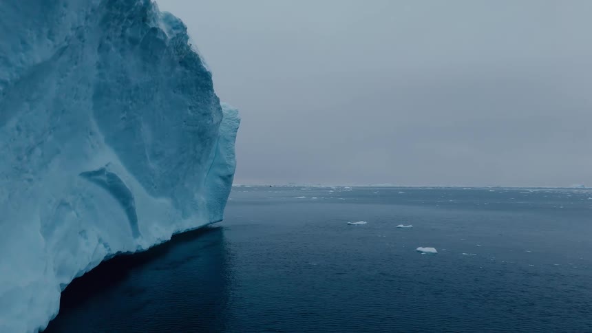 A vast dark blue ocean is shown next to the sheer blue cliff face of a massive glacier under a cloudy overcast cold gray sky