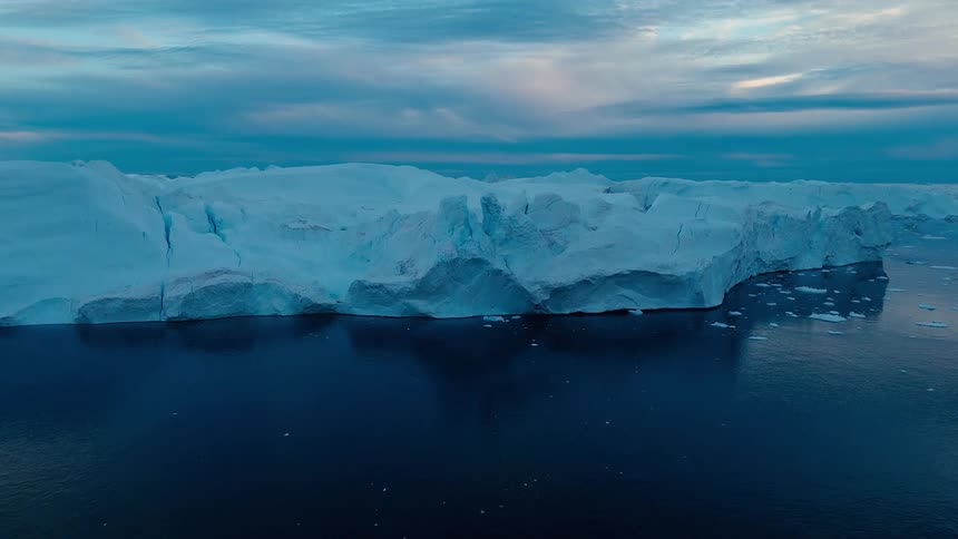 The video is an extreme long shot of a massive sheersided iceberg floating in a dark cold body of water the iceberg is a brilliant bluewhite against a dramatic blue and overcast sky with the deep water reflecting the low light