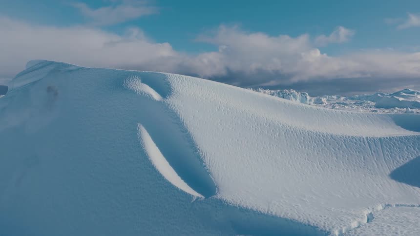 A large expanse of white ice chunks and snow drifts a massive smooth structure dominates the middle ground the terrain extends far toward a dark horizon line and white clouds drift across a blue sky