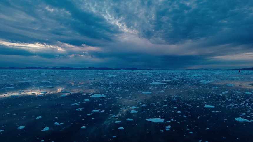 The video is a sweeping wide shot over a dark calm body of water dotted with numerous small ice floes and larger icebergs above the reflective surface the sky is dramatic with a combination of dark blue slightly ominous clouds and patches of b