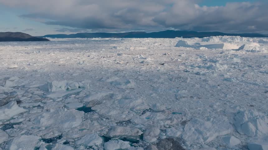 The video is a sweeping aerial shot of a vast field of ice floes and icebergs floating on the water in the background there are dark craggy hills and mountains under a cloudy sky