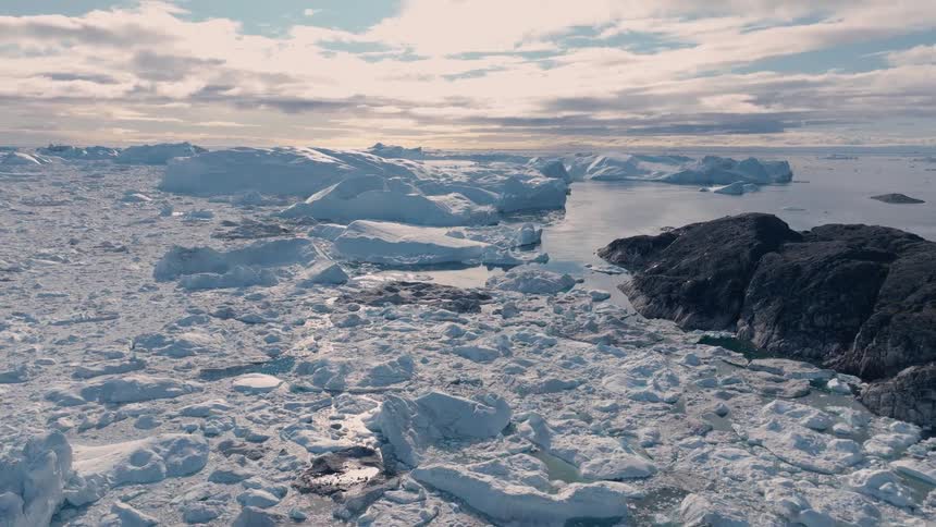 A jumbled sheet of sea ice and slush spreads toward dark rocky shorelines with small melt pools glinting in the light
