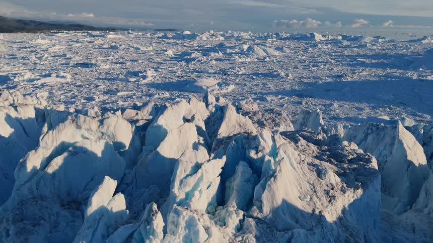 Terrain shows numerous ice peaks and deep cracks a white surface extends into the distance a horizon line separates the land from the overhead air and light falls across the broken surface