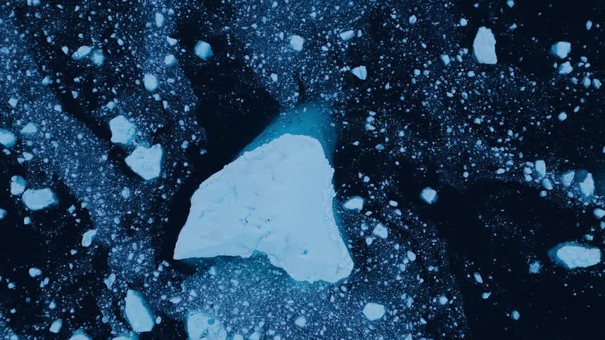 The video is a highangle extreme closeup of ice fragments and a larger piece of ice floating in deep dark blue water the water is almost black and creates a strong contrast with the bright pale blue and white ice that appears to be melting a