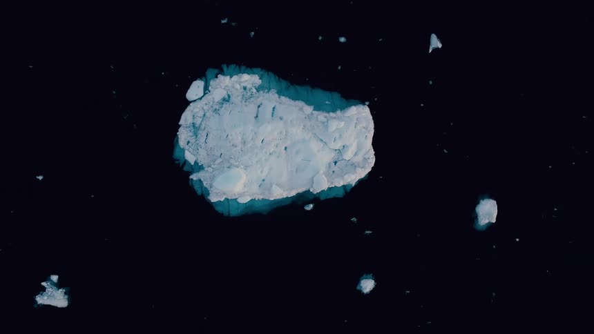 A large textured chunk of white and blue sea ice shrinking into the dark water as the camera slowly pulls back to reveal it surrounded by smaller ice fragments