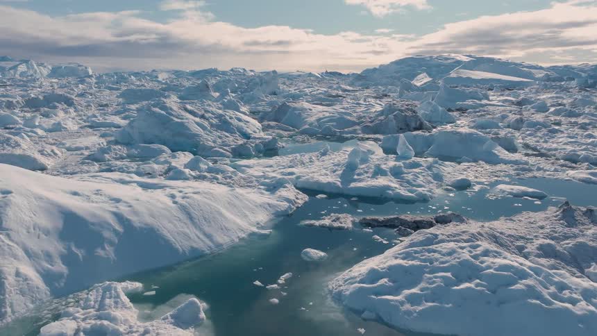 A bright polar scene shows turquoise meltwater ponds among rounded ice hummocks under soft clouds and low sun