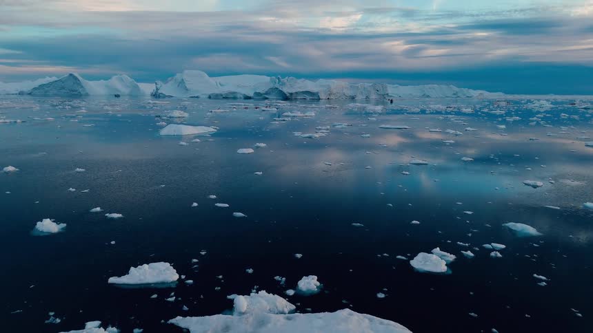The video is a wide elevated shot of a large field of small icebergs and sea ice in a deep blue body of water in the background there is a wall of massive pale blue glaciers under an overcast sky with streaks of pink and blue