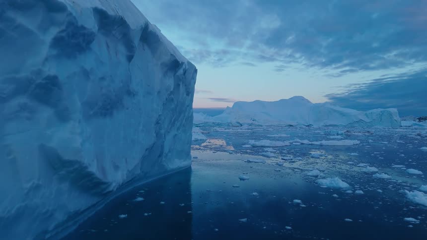 A towering iceberg rises beside a glassy icestrewn sea in cool light