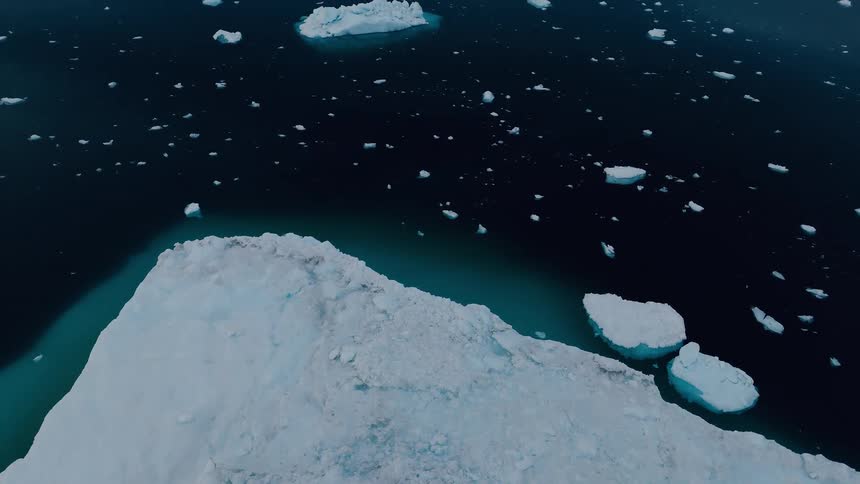 A camera moves away from the edge of a vast white ice block the frozen surface gives way to a dark blue water body numerous small pieces of ice float in the sea and the overall scene is from a high perspective
