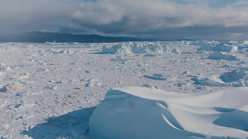 A vast field of fragmented ice stretches across calm water with distant dark mountains beneath heavy clouds