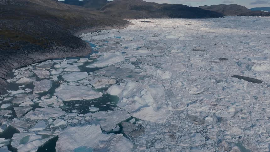 An overhead camera shot moves slowly downward and forward over a massive field of floating ice chunks and water bordered by a steep rock mountain range under a partly cloudy sky