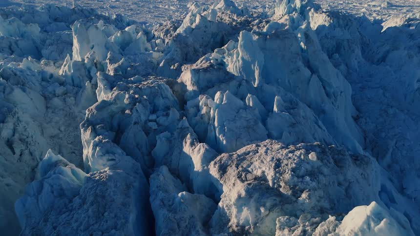 An aerial view reveals the vast rugged expanse of a glacier with deep blue crevasses and jagged ice peaks highlighted by sunlight