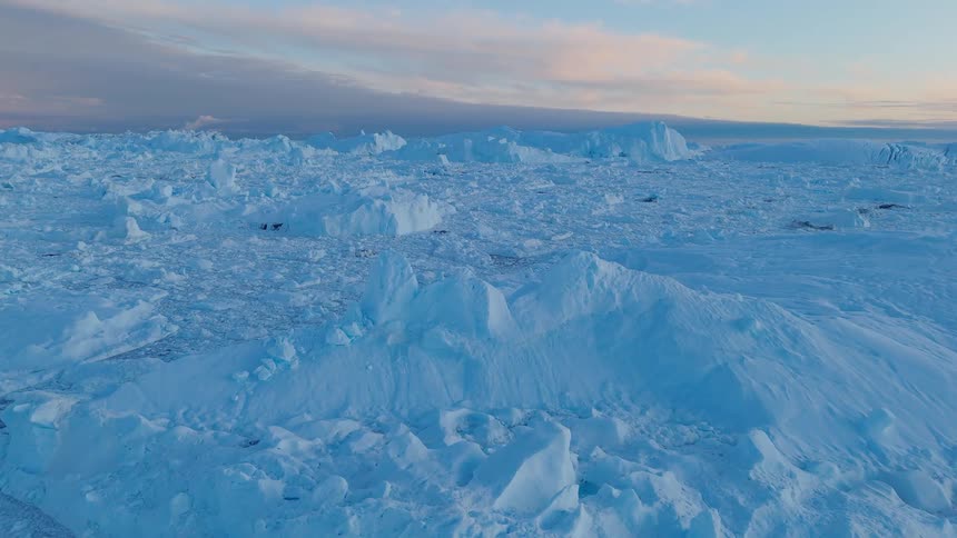 The video is an aerial shot showing a vast bright blue and white landscape of broken sea ice and icebergs the terrain is rugged and uneven consisting of many jagged chunks of ice and stretches out to a darker horizon under a pale sky