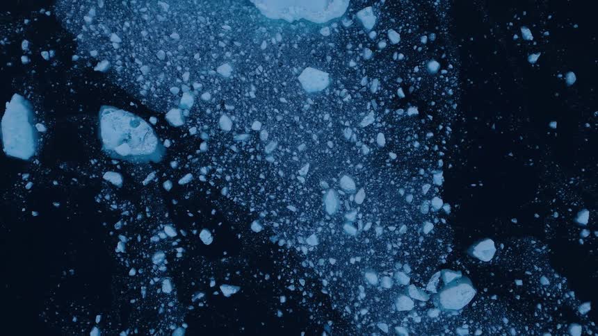 An overhead aerial view shows a dark blue body of water filled with thousands of small bright blue ice chunks and chunks of sea ice scattered across the surface