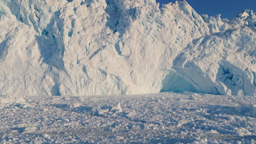 The video shows a massive jagged wall of white glacier or ice cliff rising dramatically from a foreground composed of broken heavily textured snow and ice fragments the scene is brightly lit by the sun highlighting the bluetinged crevices in