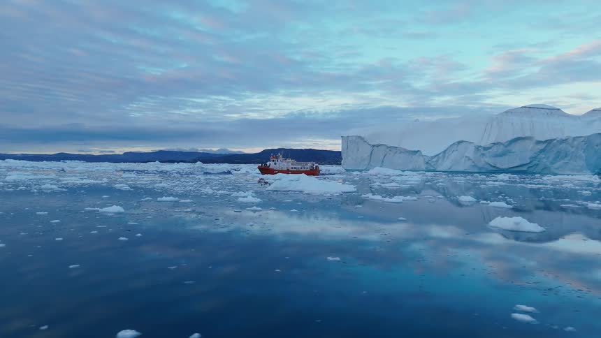 A bright orange boat navigates through a serene icy sea filled with floating icebergs and floes with a massive glacier wall and distant mountains visible under a dramatic cloudy sky at dusk or dawn