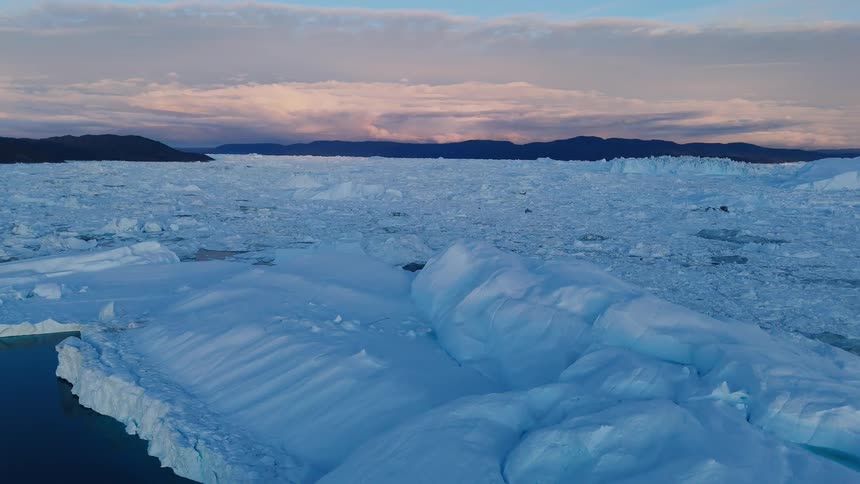 A camera moves forward across a surface of countless floating ice pieces large white icebergs border the water dark land masses lie on the horizon and a soft glow fills the cloudcovered sky