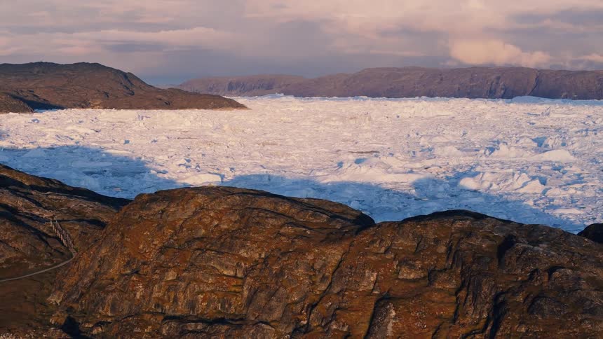 A foreground of dark rough rock contrasts with a massive field of cracked white ice the ice extends back toward a ridge of dark land and the overhead air contains scattered clouds over the mountainous environment