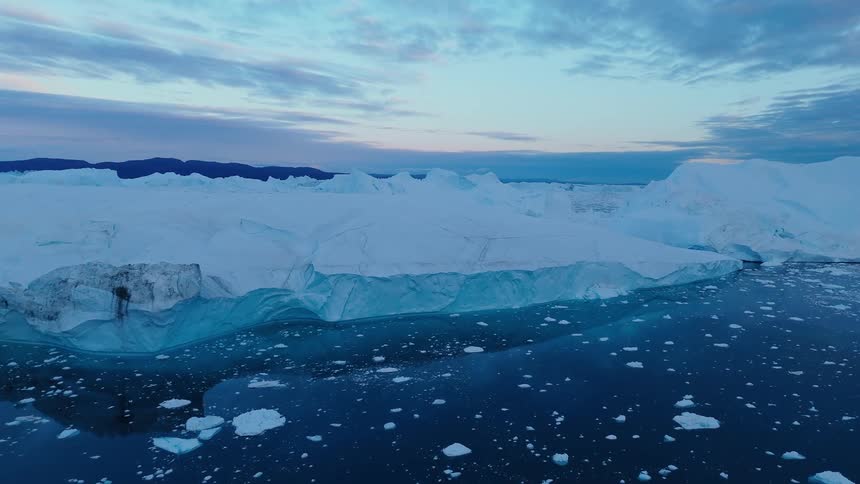 The video is a sweeping aerial shot of a vast icy and mountainous landscape at dusk it captures massive bluetinged icebergs floating in dark water under a sky painted with soft pink and blue twilight colors