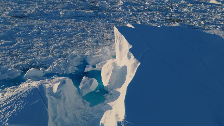 An aerial view of the frozen arctic sea shows a towering steep iceberg cliff next to glowing turquoise meltwater surrounded by flat fields of crushed ice under bright sunlight
