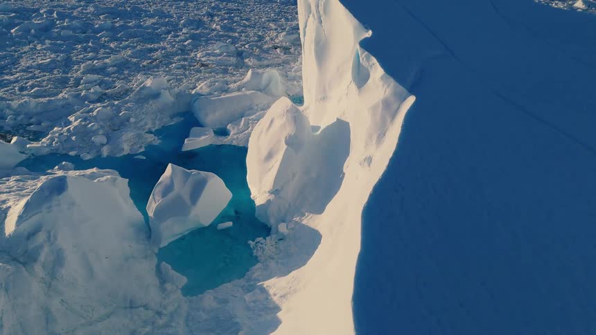 An aerial close view looks down at a massive sheer white iceberg casting a deep blue shadow with patches of bright turquoise meltwater visible between the surrounding chunks of ice