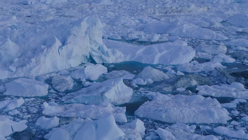 A dark rock outcrop fills the near foreground a wide area of broken white ice chunks floats in blue water the camera moves upward and over the ice surface and a pale sky hangs over the horizon