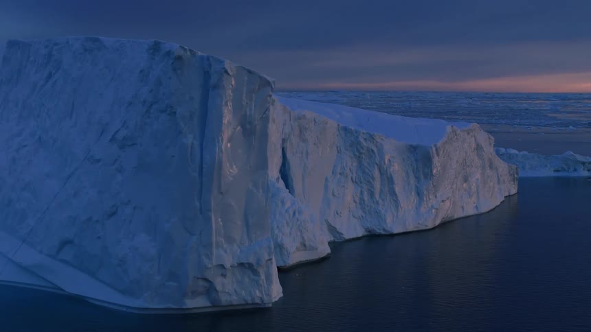 A massive wall of bluewhite ice rising from dark water with the rugged glacial structure lit by a low light source near the water line set against a dark blue and orange sky