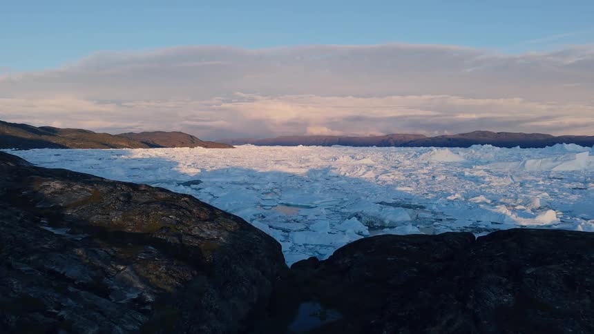 The video is a sweeping drone shot of a rocky fjordlike landscape in greenland with icebergs floating in dark blue water the foreground features large dark brown cliffs while a massive white ice sheet or glacier is visible in the distance un