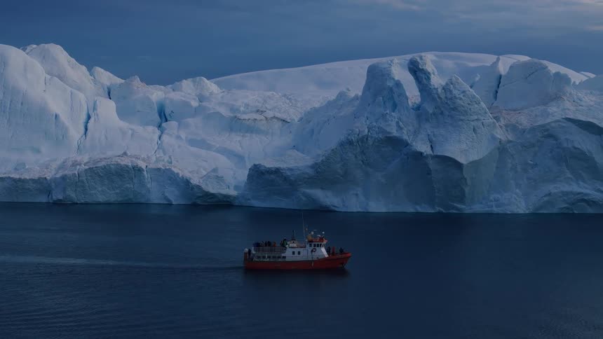 The video is a wide shot of a small orange and white boat sailing in dark blue waters in the background there is a massive towering white glacier with sharp peaks under a pale blue sky