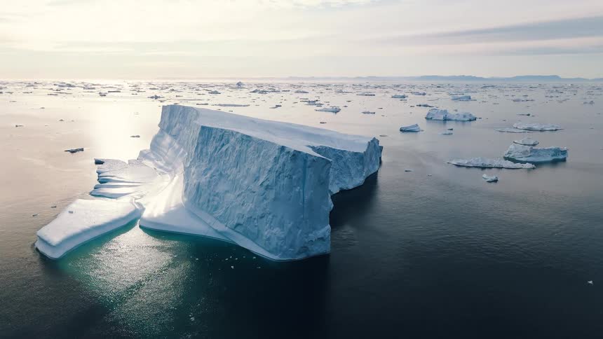 An aerial shot shows a massive angular iceberg floating in a sea dotted with smaller ice chunks dramatically reflecting the blue and green tones of the water the sky above is bright and hazy suggesting either midday sun or a pale twilight