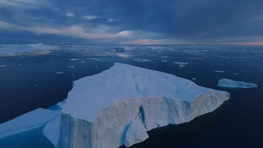An aerial view shows massive white icebergs floating in a dark blue cold ocean under a dramatic cloudy sky at twilight or sunset