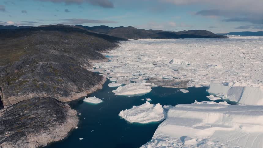 A wide expanse of white ice fragments and large icebergs meets the edge of a dark rocky coastline filling a deep blue fjord under a partly cloudy sky