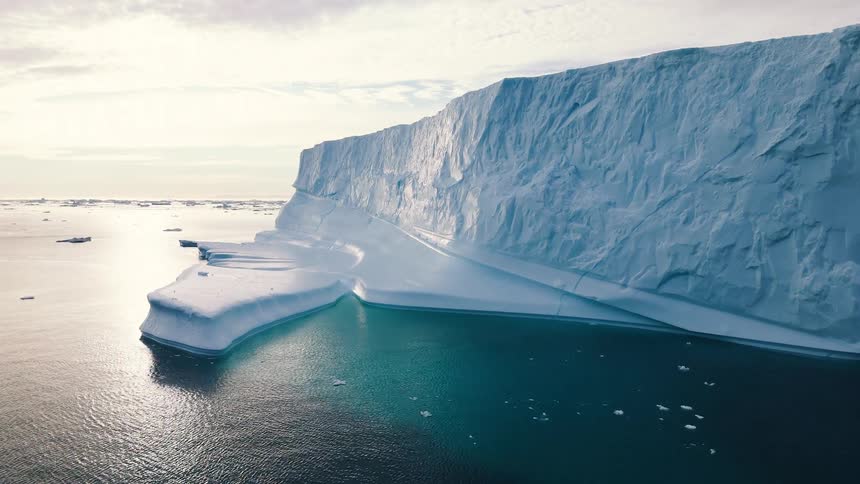 The video is an extreme long shot of a small sailboat with a red sail passing in front of a massive sheersided blueandwhite iceberg the boat is dwarfed by the glacial ice emphasizing the scale of the icy open water landscape under a bright