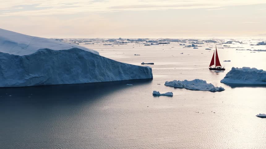 The video captures an expansive arctic or antarctic scene dominated by massive icebergs floating in dark water with a hazy sky in the backgrounda small sailboat with a vibrant red sail moves past the ice providing a striking contrast agai