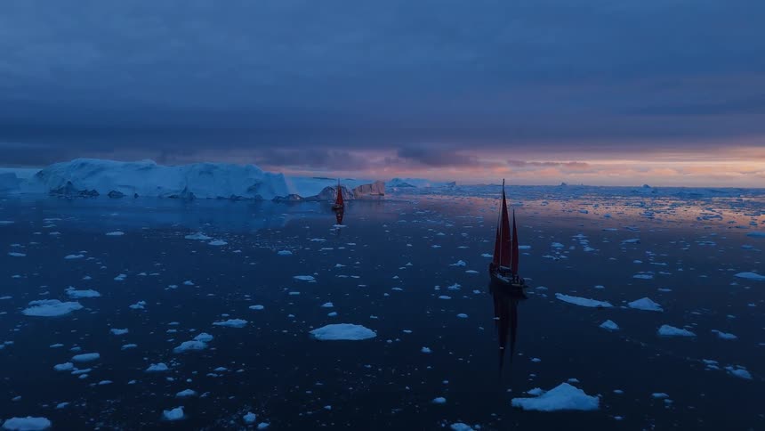 The video is a wide elevated shot of a sailboat navigating through a sea dotted with ice floes and large icebergsthe scene is captured during twilight giving the water a deep blue color and the horizon a soft orange and purple glow