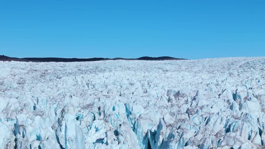 This is a vast static shot of a massive bright blue and white glacier with deeply crevassed jagged ice formations in the foreground above the glacier the horizon is marked by dark distant mountains under a clear brilliant blue sky
