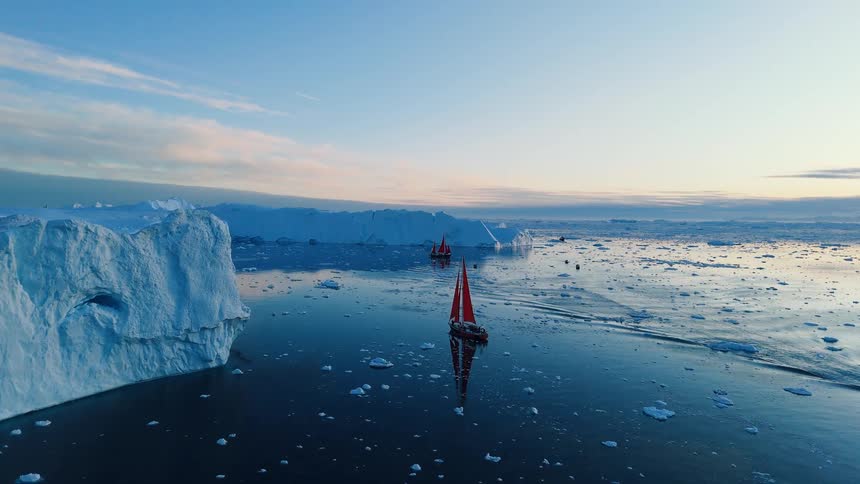An aerial shot shows a single redsailed sailboat and a small motorboat cruising across a dark blue icechoked sea a massive white and blue glacier towers over the scene on the left with the horizon filled with countless smaller icebergs an