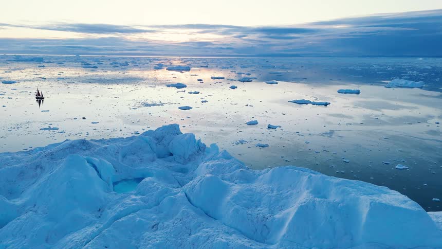 An aerial view of the cold arctic ocean shows numerous ice floes floating beyond a large rocky iceberg in the foreground with a tiny sailboat visible on the water under a soft twilight sky