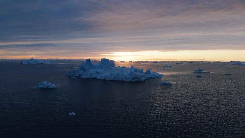 An aerial camera shot moves over a dark water body scattered with large and small icebergs as the low sun creates a streak of light reflection across the water setting against a colorful cloudy sky