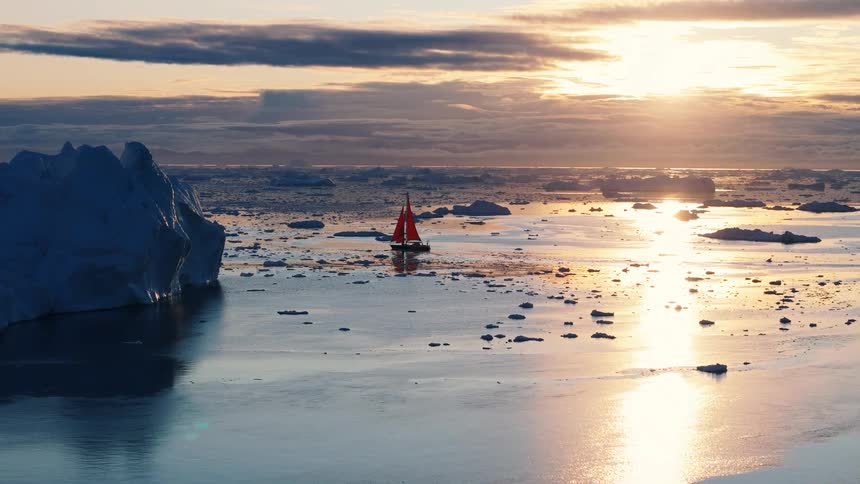 Aerial view of a sailboat with red sail moving through icy waters at sunset